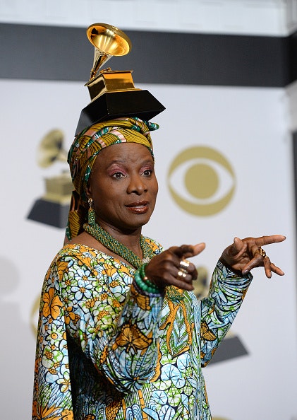 A woman in African attire points her fingers to the room as she poses for a photo with a gold statue of a gramophone balancing on her head.