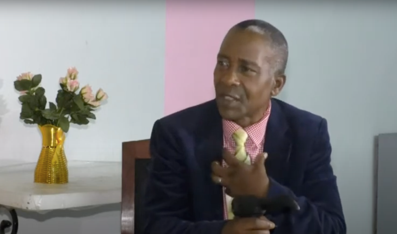 An elderly man sits in a chair with one hand on a walking stick and another gesturing. He is distinguished, in a jacket and tie.