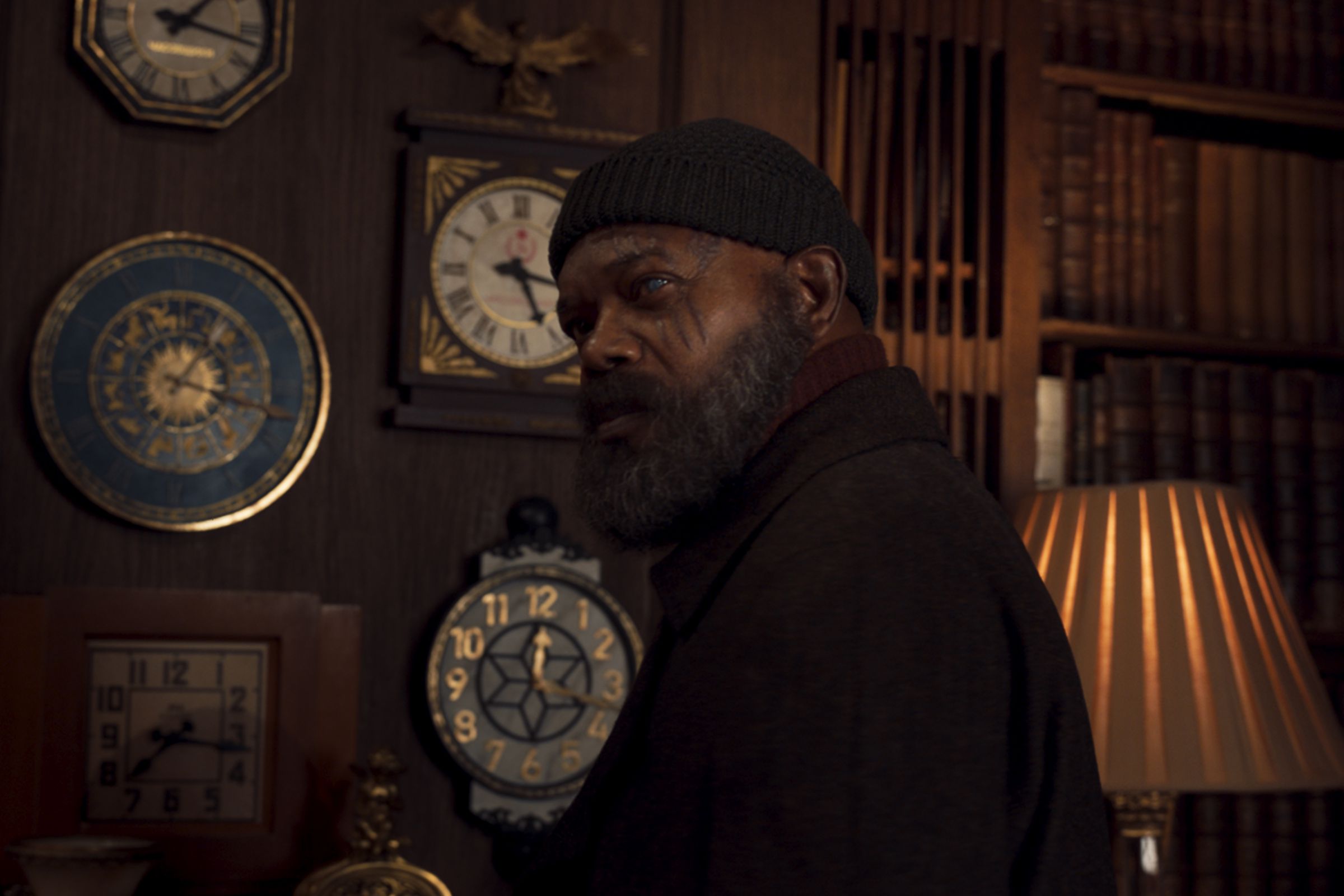 A bald, bearded man wearing a black beanie and black turtleneck, and standing in front of a wall lined with books and clocks.