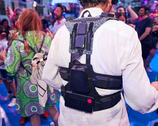A person wearing a white shirt stands in a crowd of people dancing at a party. The person is wearing a black device called a haptic suit that wraps around the shoulders and the waist like a backpack.