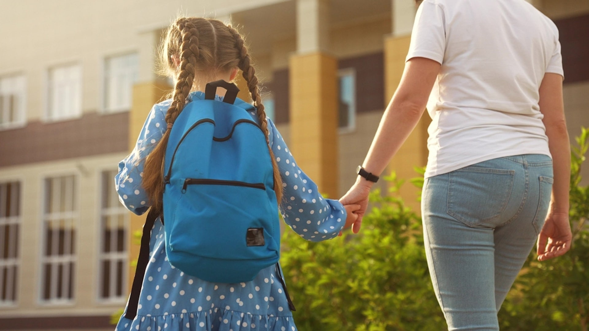 A mother leads her child by the hand toward a building. The child is wearing a backpack.