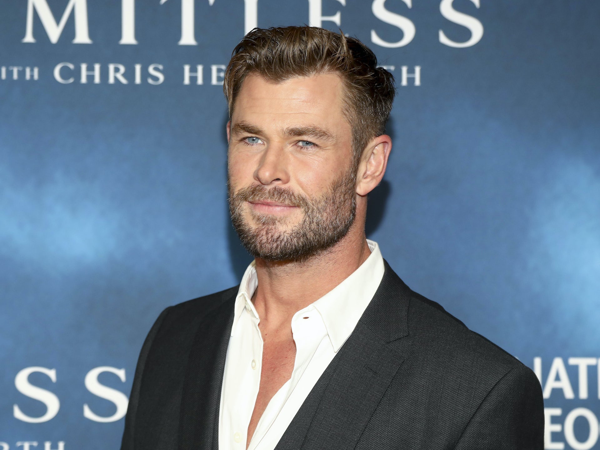 Actor Chris Hemsworth wearing a dark suit jacket and white shirt with open collar in front of a blue backdrop that reads 'Limitless with Chris Hemsworth'