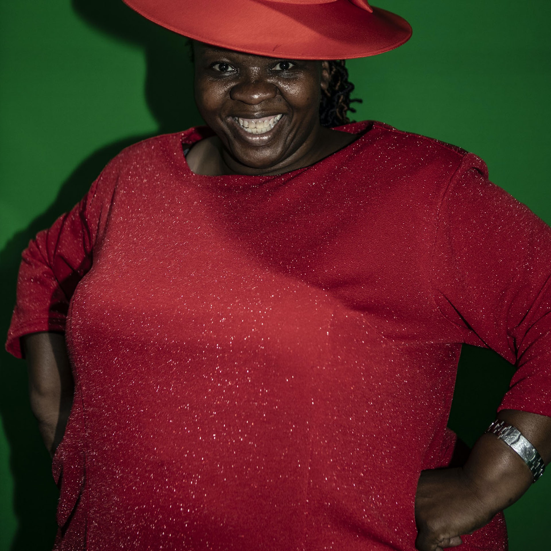 A woman smiles, hands on her hips in a shimmering red gown and hat.