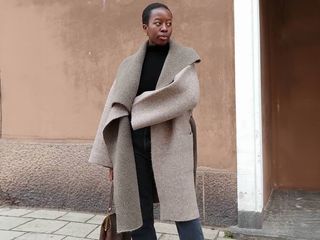 European fashion influencer Sylvie Mus poses on a Paris sidewalk with an oversize neutral coat, black mock neck top, jeans, and a brown bag