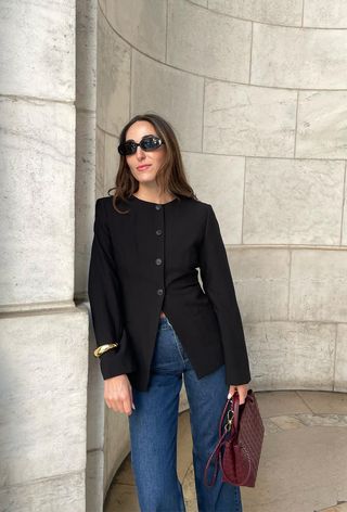 one of the best chunky cuff bracelets is shown in a photo of a woman standing outside wearing black sunglasses with a black blazer, jeans, a red handbag, and a gold cuff bracelet
