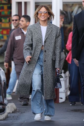 Jennifer Lopez wears a gray long wool coat, cropped white tee, baggy jeans, white sneakers, and a black Hermes bag.