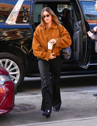 Hailey Bieber wears a brown suede jacket, The Row Pim bag, black trousers, and black loafers.