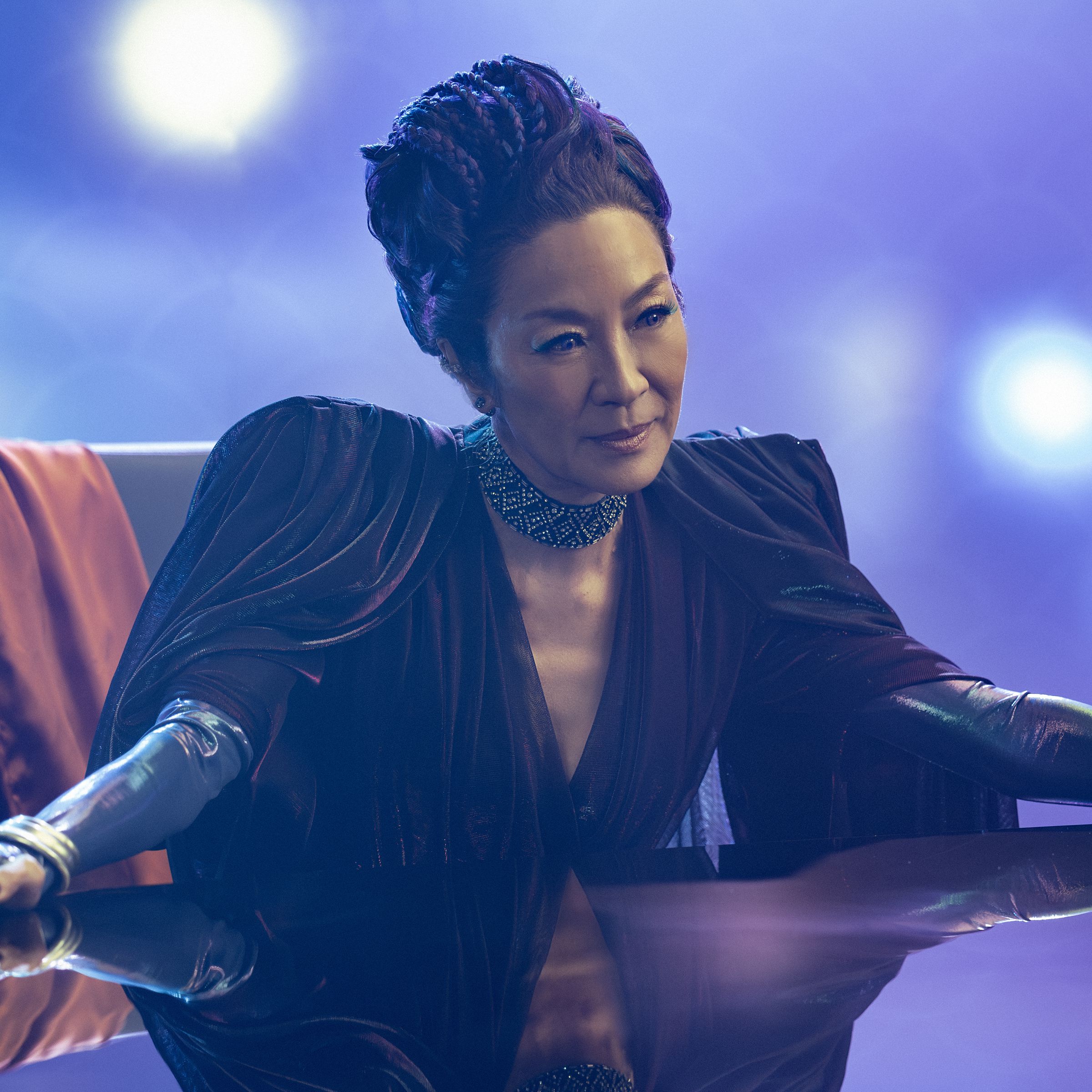 A woman in an elaborate gown with pointed shoulder pads sitting at a large desk in a chair with her arms spread out in front of her.