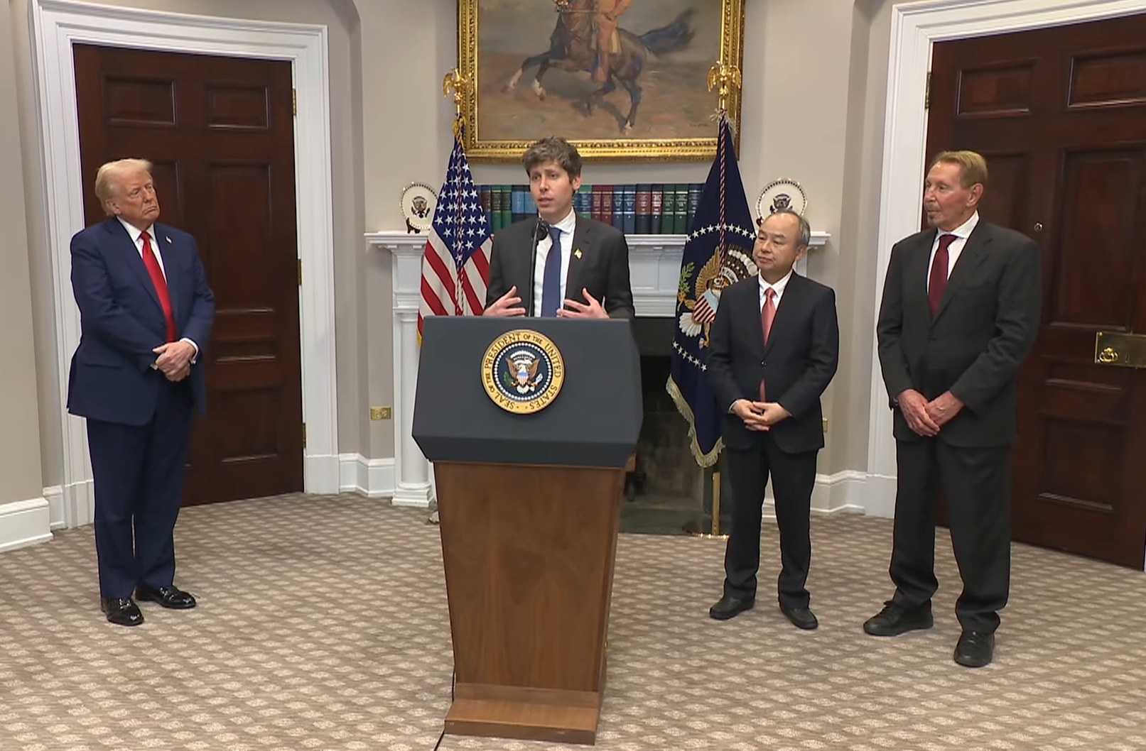 Donald Trump standing off to the side while OpenAI CEO Sam Altman speaks behind a lectern at the White House.