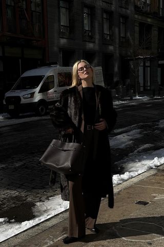 dark brown outfit idea shown in a photo of a woman standing on the sidewalk in New York wearing brown glasses with a dark brown fur coat layered over a brown blazer and black boatneck blouse styled with brown trousers, brown ankle boots, and a brown leather tote bag
