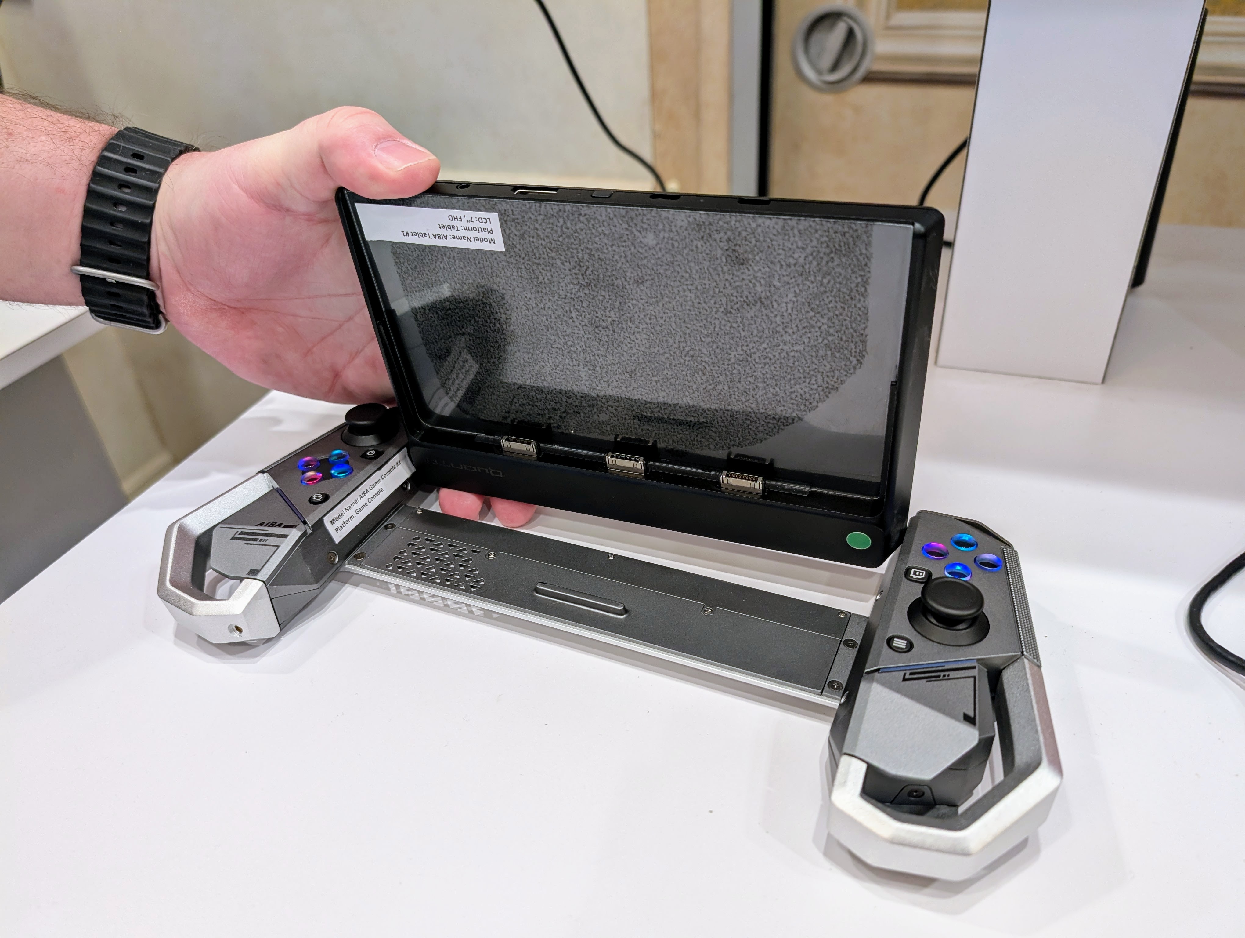 A handheld gaming controls set in a metal bar that spans a tablet screen which is lifted to show connectors on the underside.