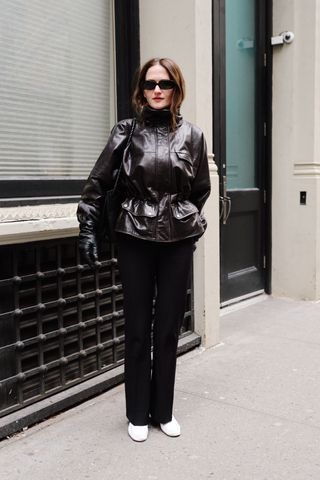Eliza Huber wearing a brown leather jacket, black trousers, white loafers, and black sunglasses while carrying a black Bottega Veneta Ciao Ciao bag outside of the Kallmeyer show in Tribeca.