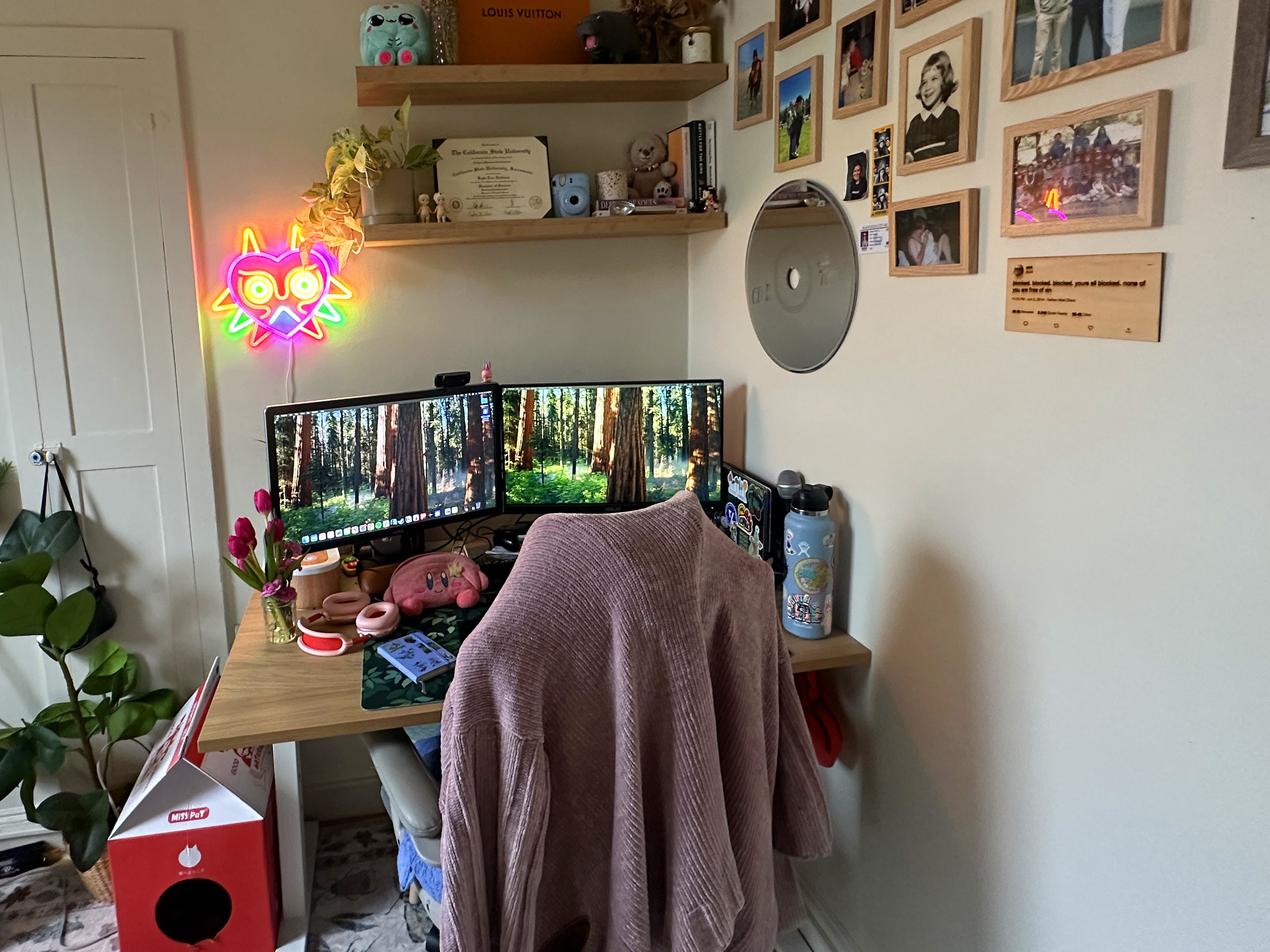 Desk in a corner with monitors, photos on the wall, and a chair with a sweater draped over it.