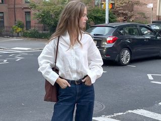 Scandinavian influencer and creative Clara Dyrhauge poses at a NYC crosswalk wearing a white linen button-down shirt, a brown leather tote bag, black belt, and dark-wash jeans.