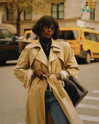 New-York-based fashion influencer @lefevrediary wearing a classic trench coat with jeans on the street in Manhattan, with yellow cabs in the background.