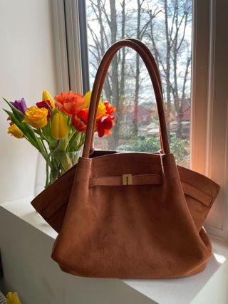 JW Pei Hana Tote Bag in tan faux suede sits on window sill in front of a bunch of flowers.
