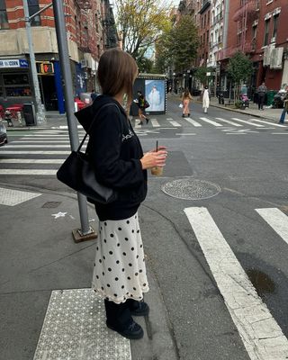 Poppy Almond wears a white and black polka dot skirt with a hoodie and a coffee in hand.