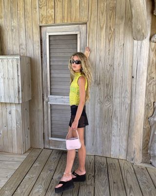 Emili Sindlev wears a yellow top, black miniskirt, and black platform flip flops.