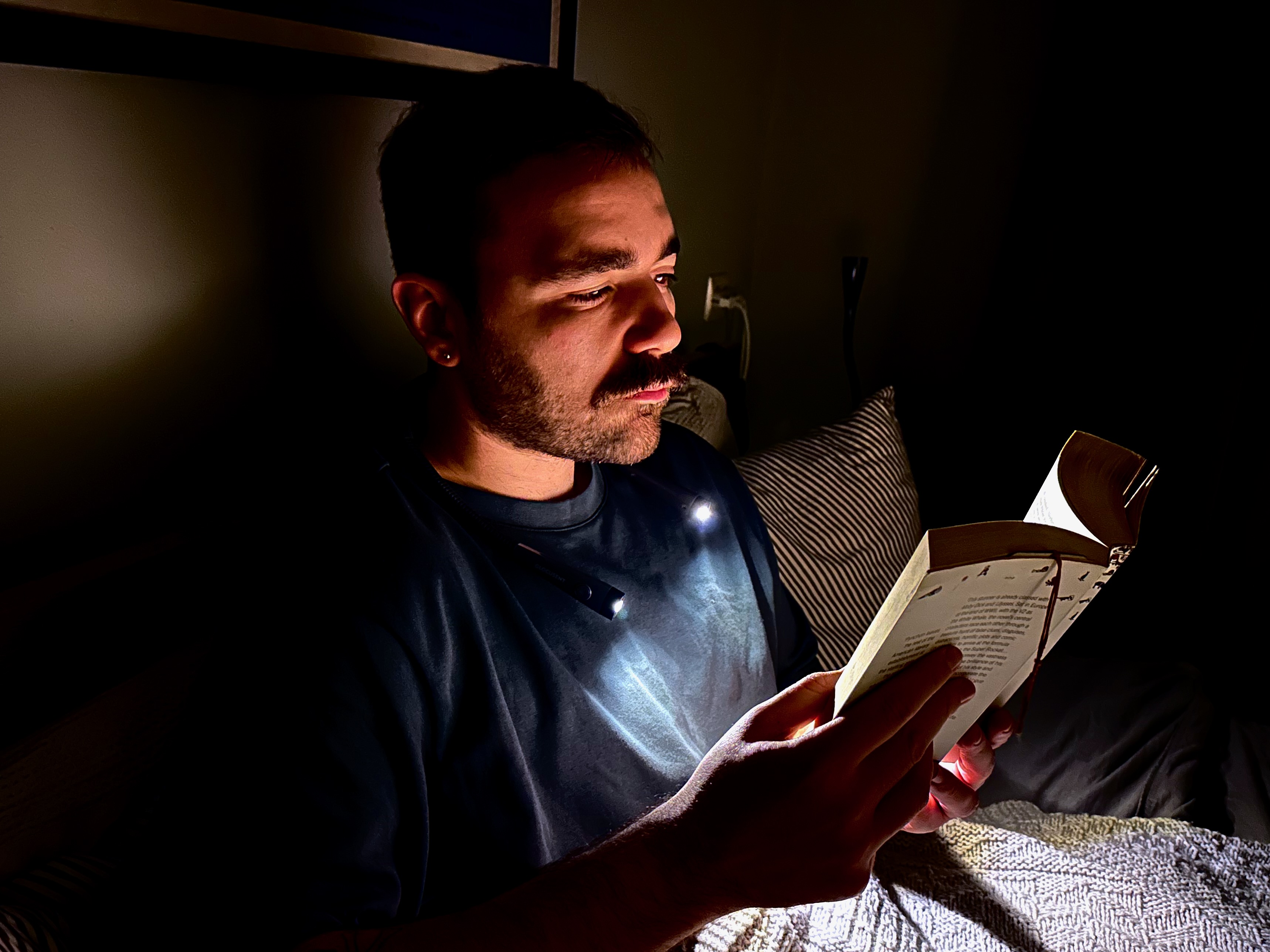 A man in a dark bedroom wears a neck lamp that illuminates just his face and the book he’s holding while sitting upright in bed.