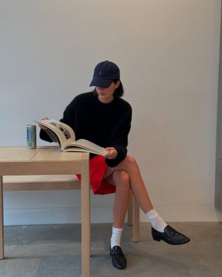 @kendalljenner wears a red knee skirt with white socks, black loafers, a navy jumper and a baseball cap