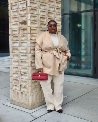 Abi wears a beige wrap coat, white top, beige pants, and a red savette bag.