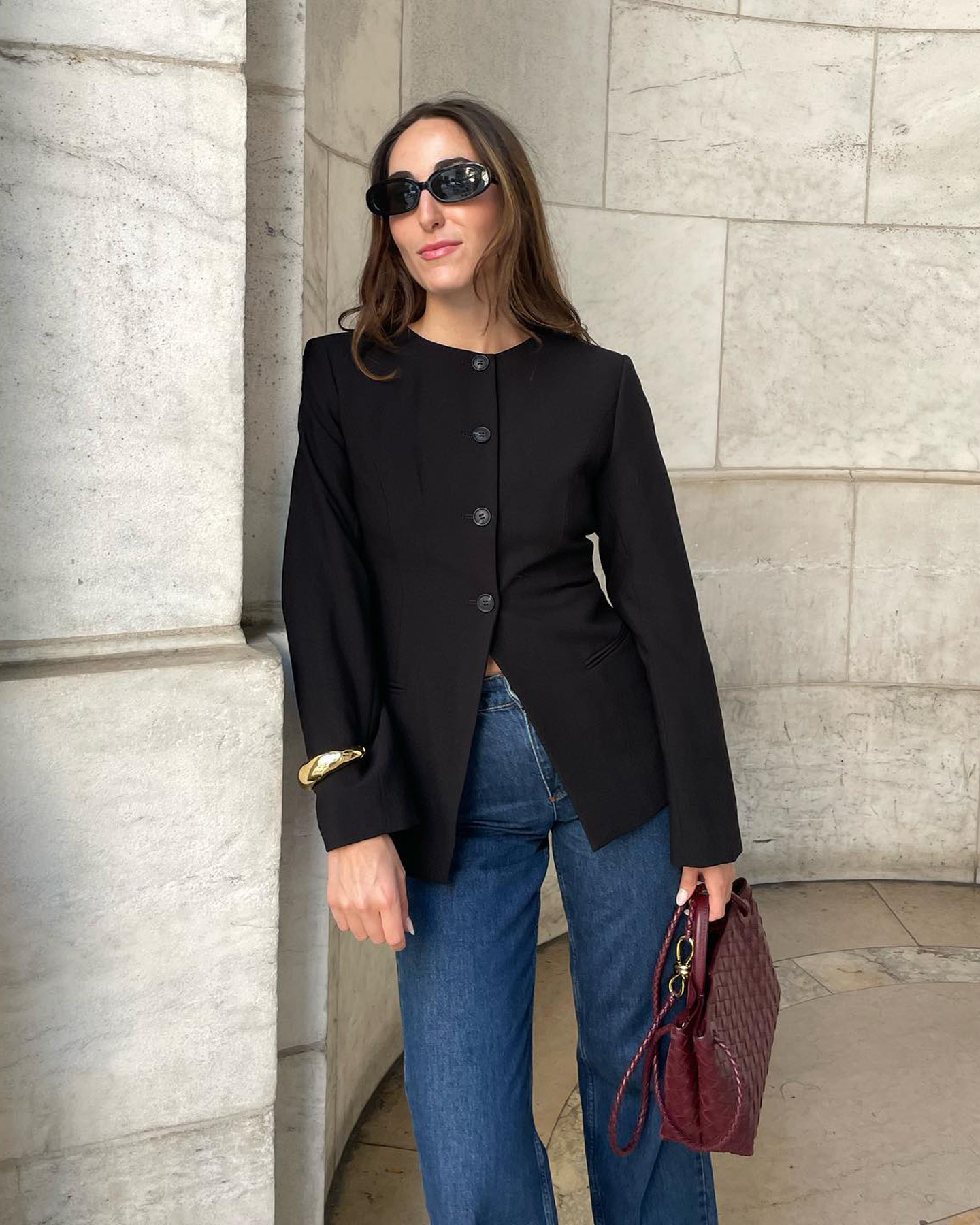 fashion editor Anna LaPlaca poses in black oversize oval sunglasses, a black Aligne button-down crew neck blazer, a gold cuff, high-waisted jeans, and burgundy woven Bottega Veneta bag