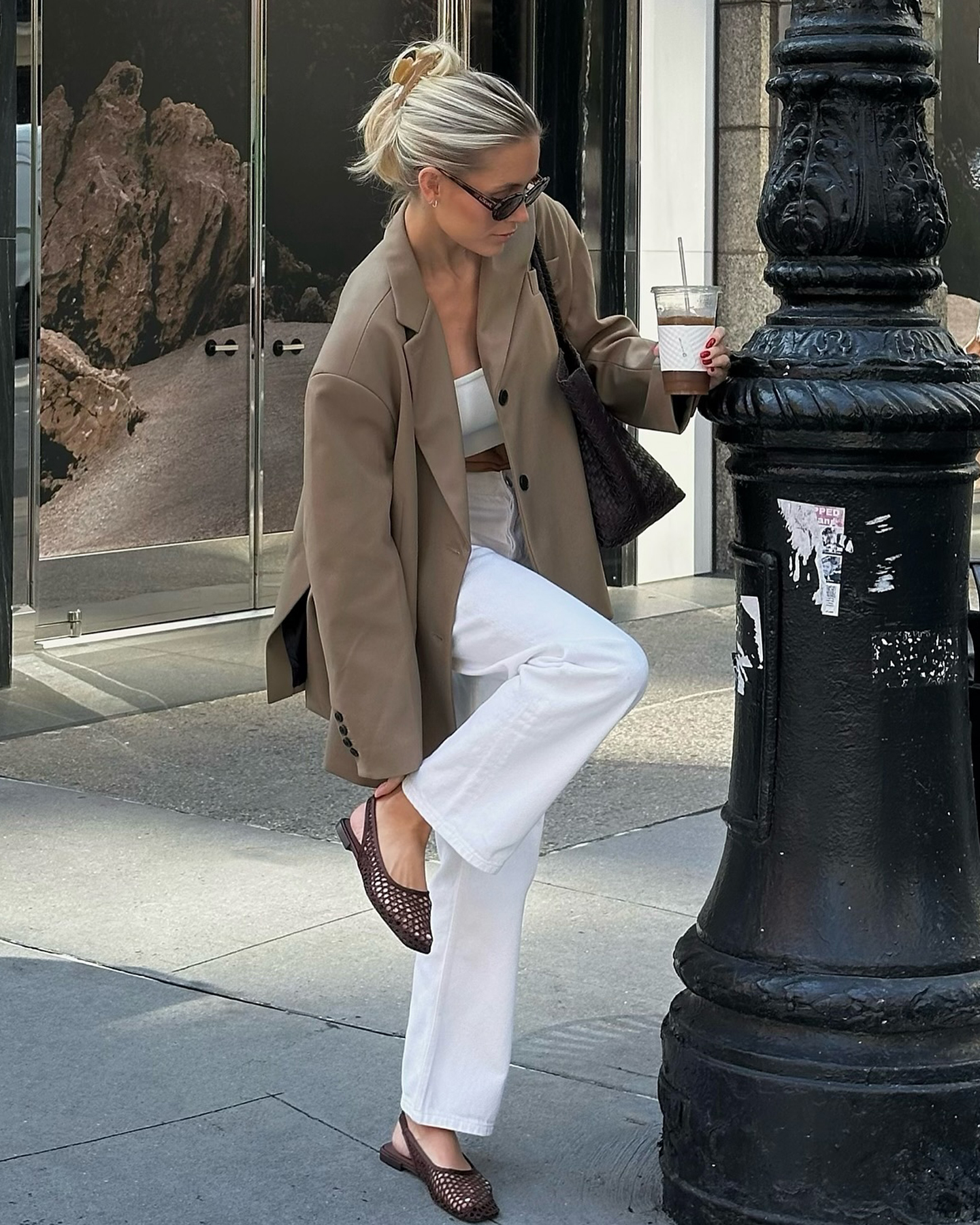 influencer Fredrika Ekerot poses on a NYC sidewalk wearing a hair claw clip, sunglasses, light brown blazer, white bandeau top, white straight-leg jeans, and brown woven slingback flats