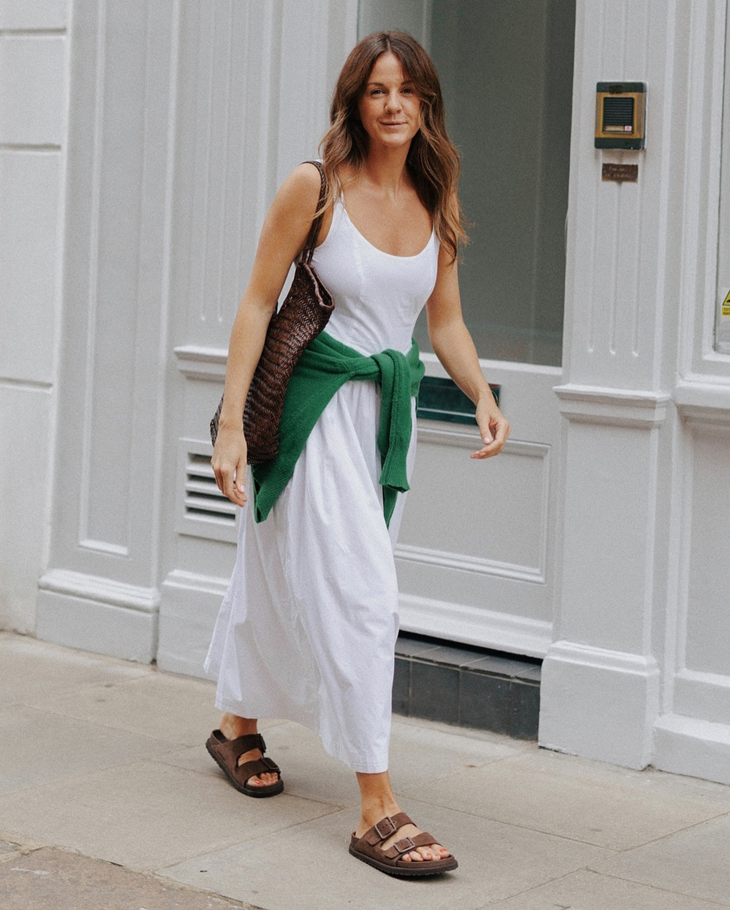 British style influencer Kim Turkington walking down a sidewalk wearing a sleeveless white poplin maxi dress, a brown woven leather tote, a green sweater tied around the waist, and brown suede Birkenstock sandals