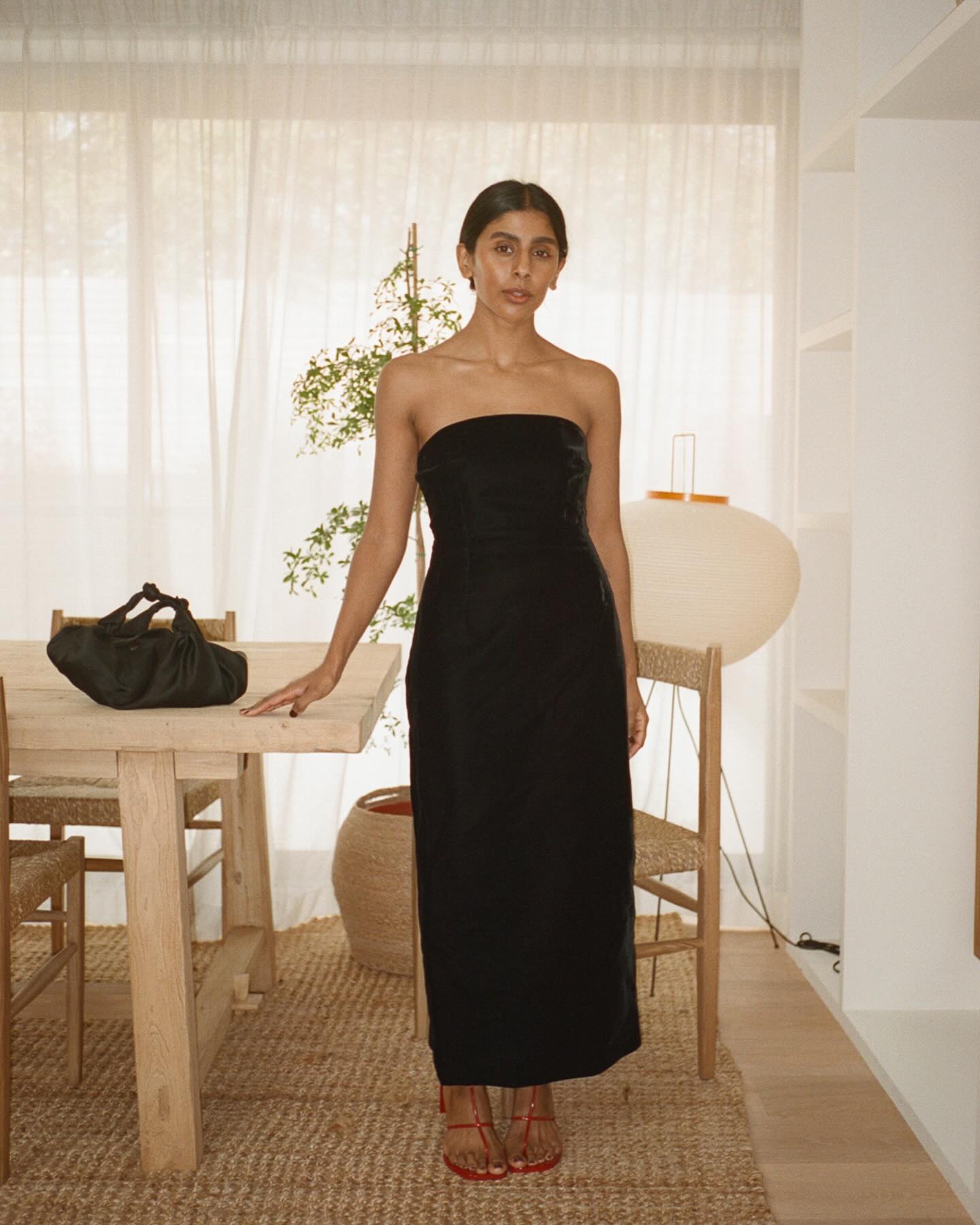 British influencer Monikh Dale poses in her London livingroom wearing a black strapless dress, red strappy barely-there heeled sandals, and a satin knotted black bag.