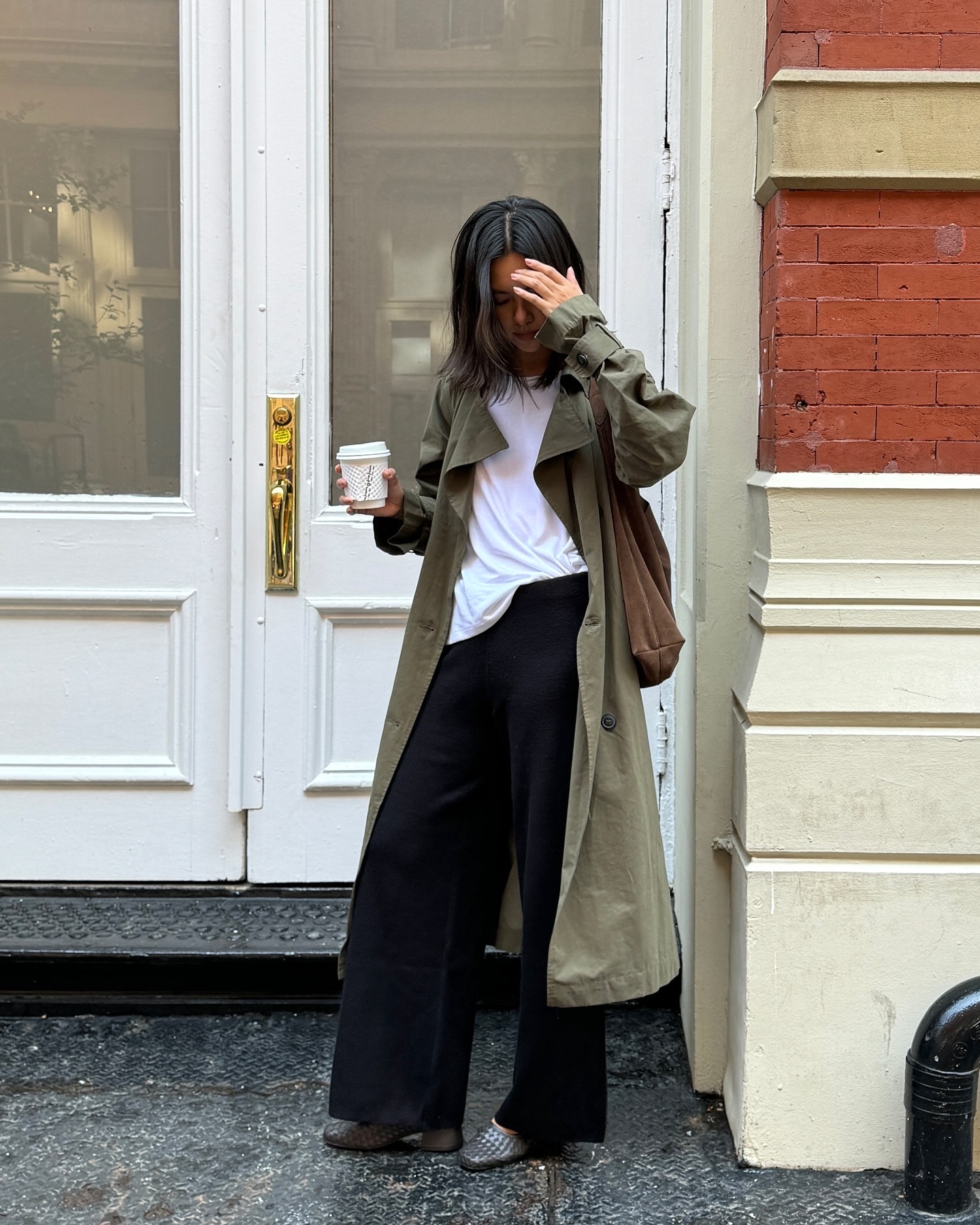 style influencer Mimi Nguyen poses on a NYC sidewalk wearing a long green trench coat, oversize white T-shirt, black wide-leg pants, and gray woven short heels