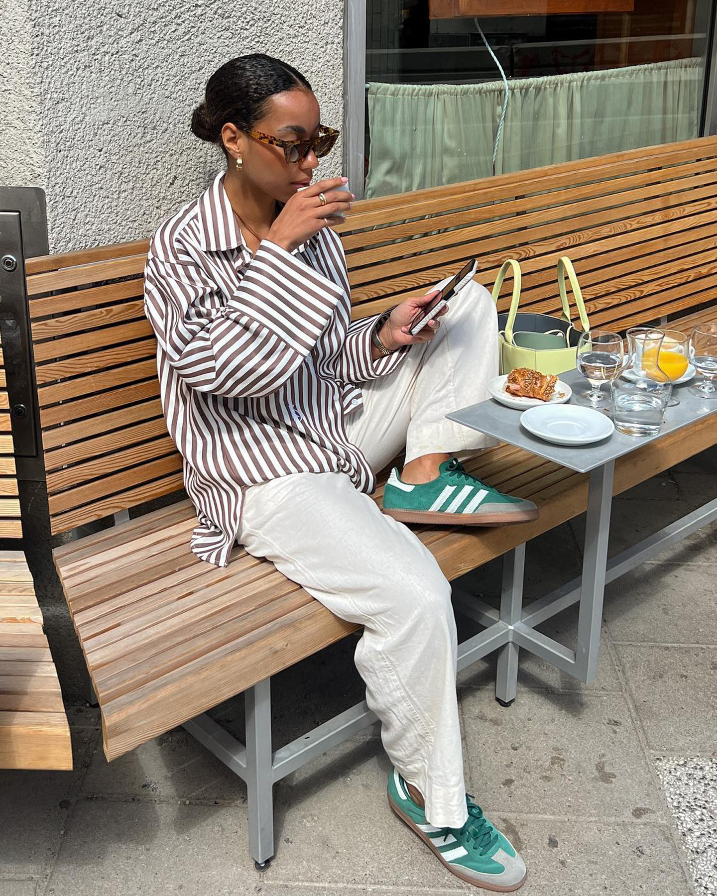 Dutch style influencer Amaka Hamelijnck sits an outdoor cafe wearing tort sunglasses, a striped button-down shirt, beige linen pants, and green Adidas Samba sneakers