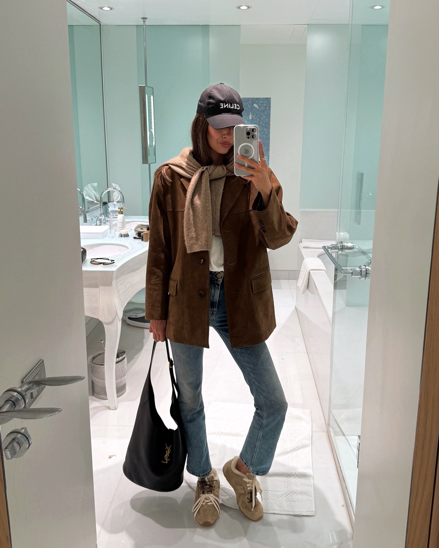 style influencer Felicia Akerstrom poses for a mirror selfie wearing a Celine baseball cap, a tan sweater tied around her shoulders, a brown suede jacket, leather tote bag, straight-leg jeans, and tan suede Miu Miu sneakers