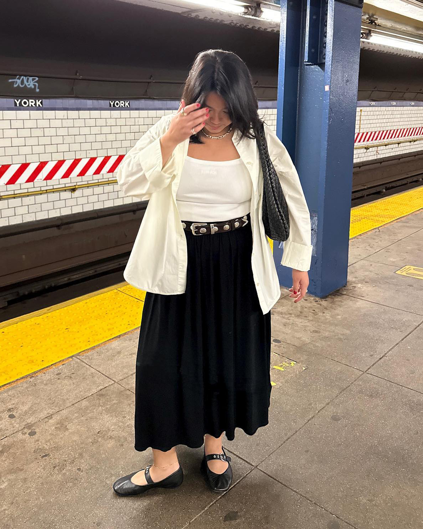mid-size style influencer Marina Torres poses on a Subway platform in NYC wearing an open white button-down shirt, a white tank top, studded black belt, full black skirt, and black Mary-Jane ballet flats