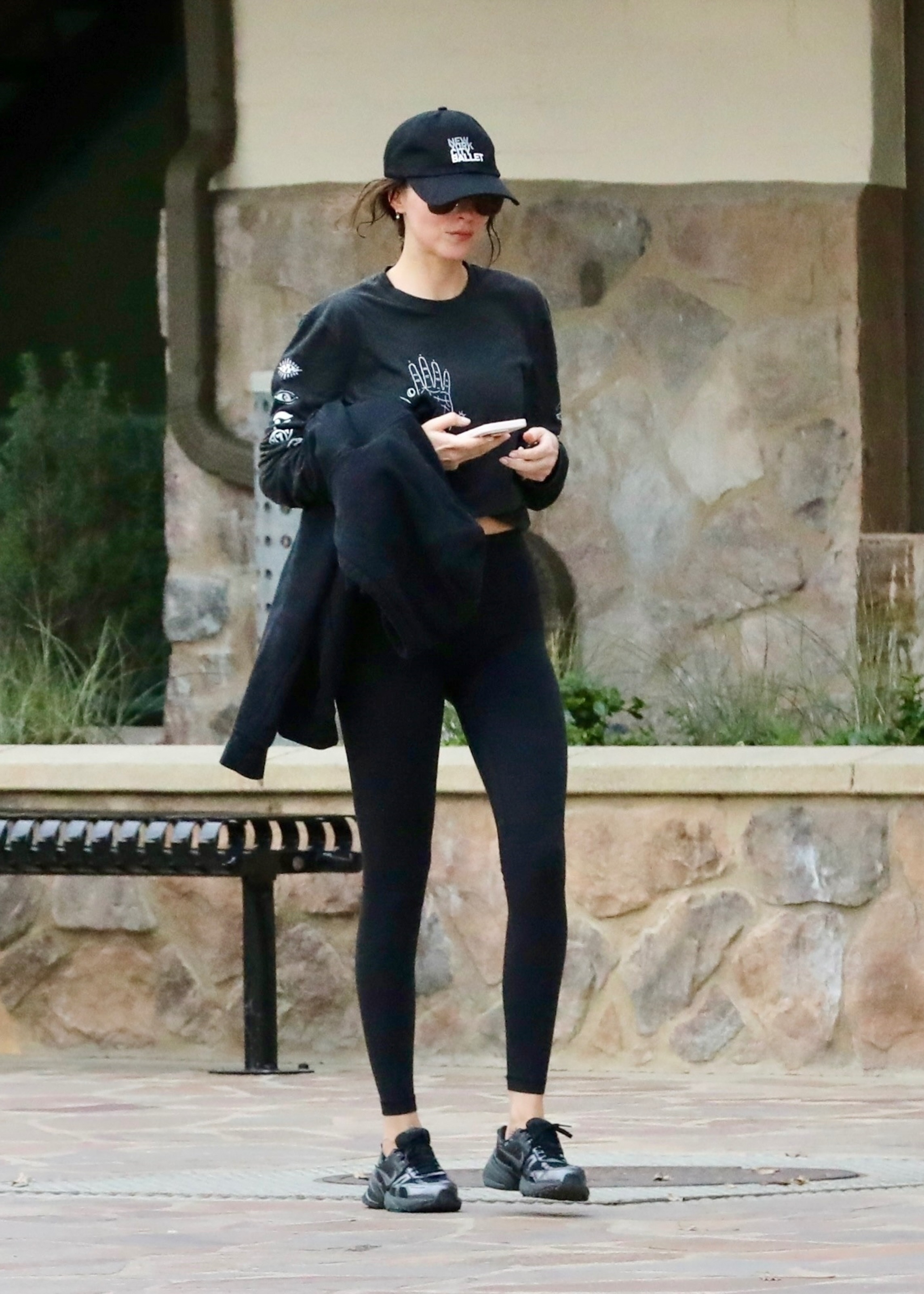 Dakota Johnson wears a black hat, black shirt, black leggings, and nike v2k black and gray sneakers.