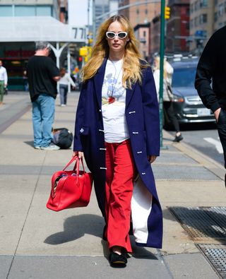 Jennifer Lawrence wears a blue car coat, white graphic tee, white sunglasses, red bag, red jeans, and black v-cut flats.