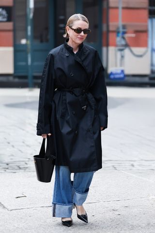 Naomi Watts wearing a Prada trench coat, jeans, heels, and a black brushed leather bucket bag in New York City.