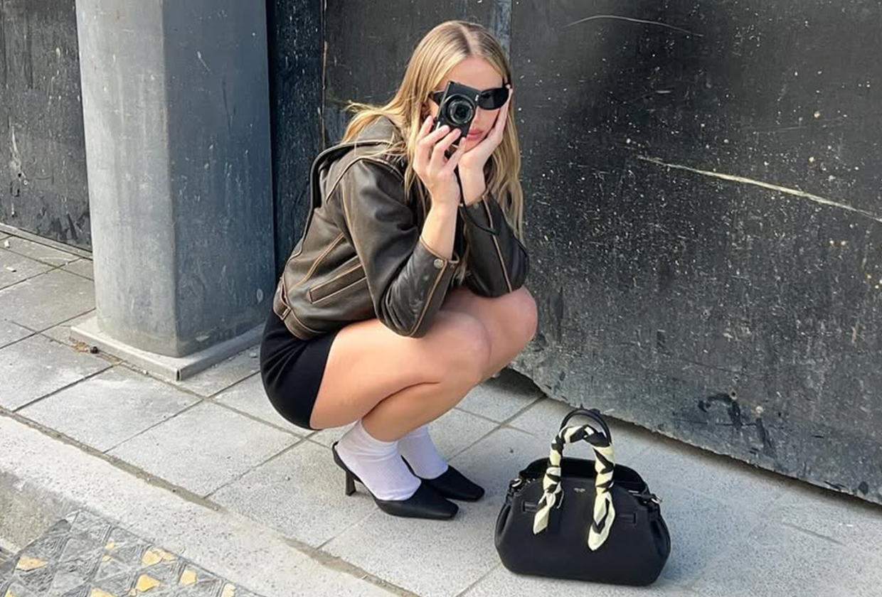 Hannah Juneva holding a black Coach bag wearing black sunglasses, a leather bomber, black shorts, white socks, and black pointed toe mules.