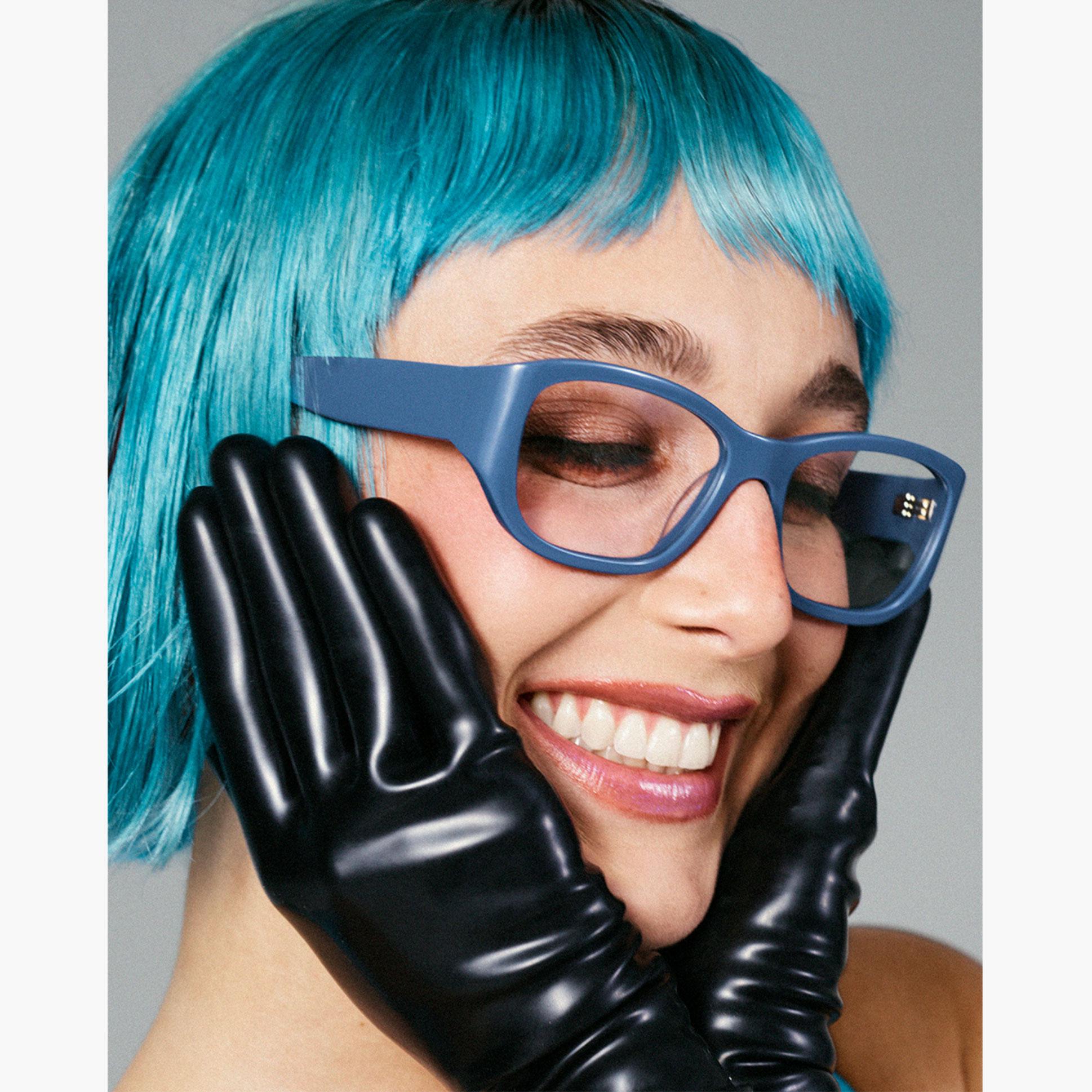 emma chamberlain wears blue acetate patty frames from her warby parker collection while wearing a blue wig and leather gloves and smiling while holding her face