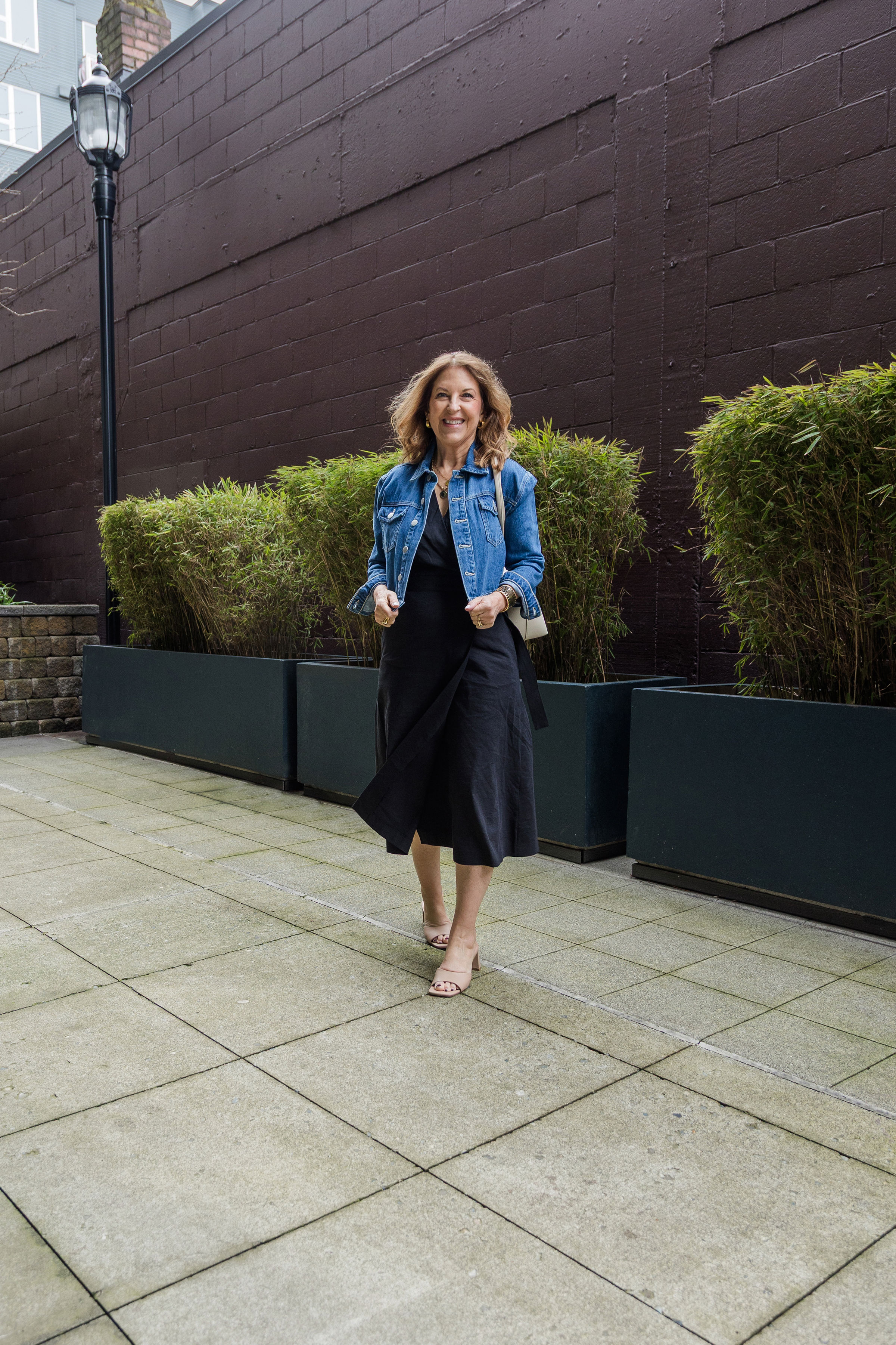 A former VIP Nordstrom stylist wearing a black midi dress, a denim jacket, and tan heeled sandals.