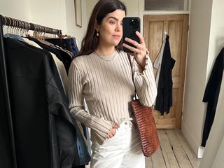 British influencer Anna Newton poses for a mirror selfie in her London home wearing a beige ribbed long-sleeve top, a brown woven tote bag, and white jeans