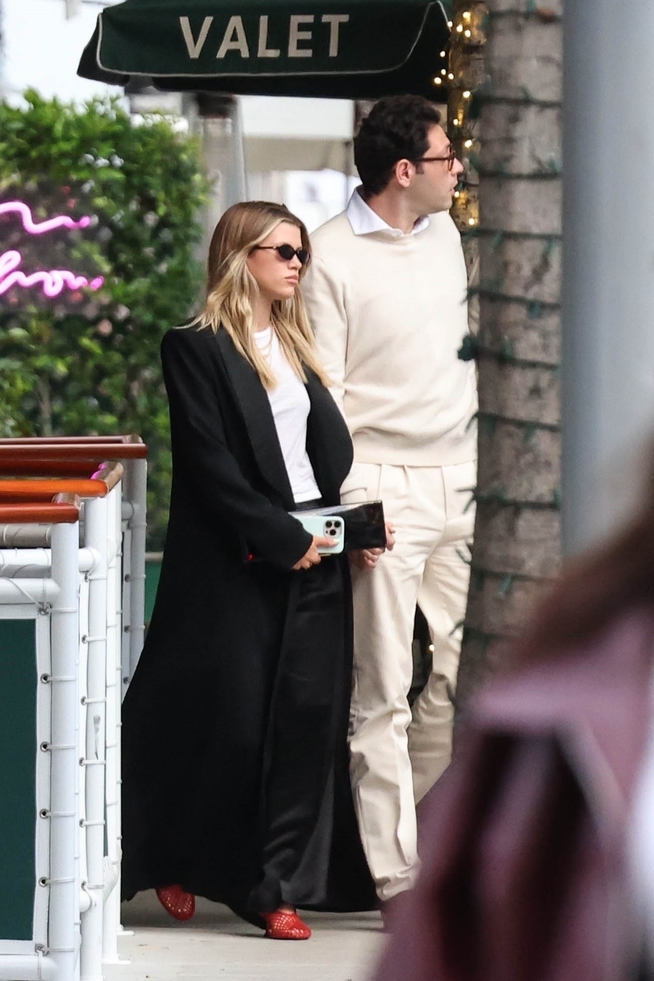Sofia Richie and Elliot Grainge walk hand in hand after dinner at Cipriani in Beverly Hills on April 26, 2025. Richie is wearing a black coat, white T-shirt, black bottoms, and red Proenza Schouler net heels.