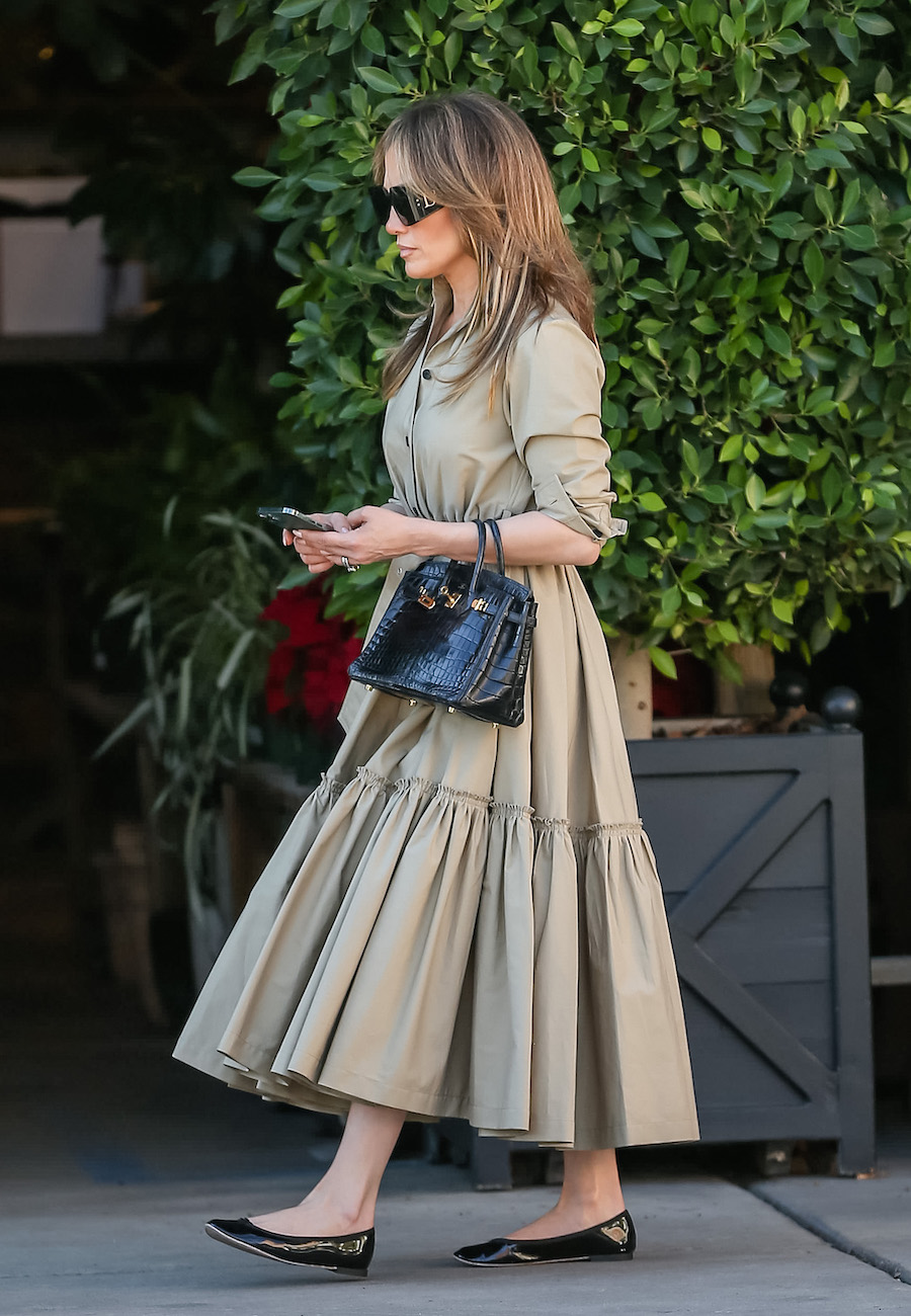 Jennifer Lopez walks with her phone in her hand wearing sunglasses, a tiered tan shirt dress, black croc Hermes bag, and black patent ballet flats