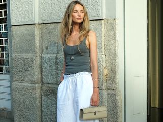 Dutch fashion influencer Anouk Yve poses on a side walk in Milan, Italy wearing a gray tank top, long silver pendant necklace, white drawstring linen pants, and a tan Savette Pochette bag