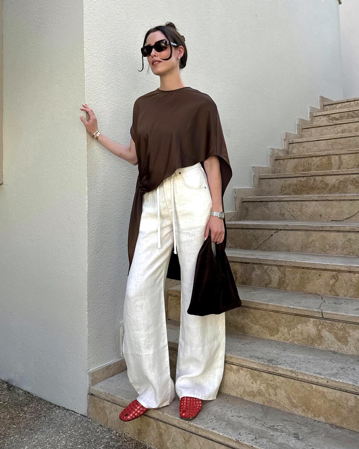style influencer Annabel Rosendahl poses on an outdoor staircase wearing a brown asymmetrical cape top, white linen pants, a black bag, and red The Row jelly flats