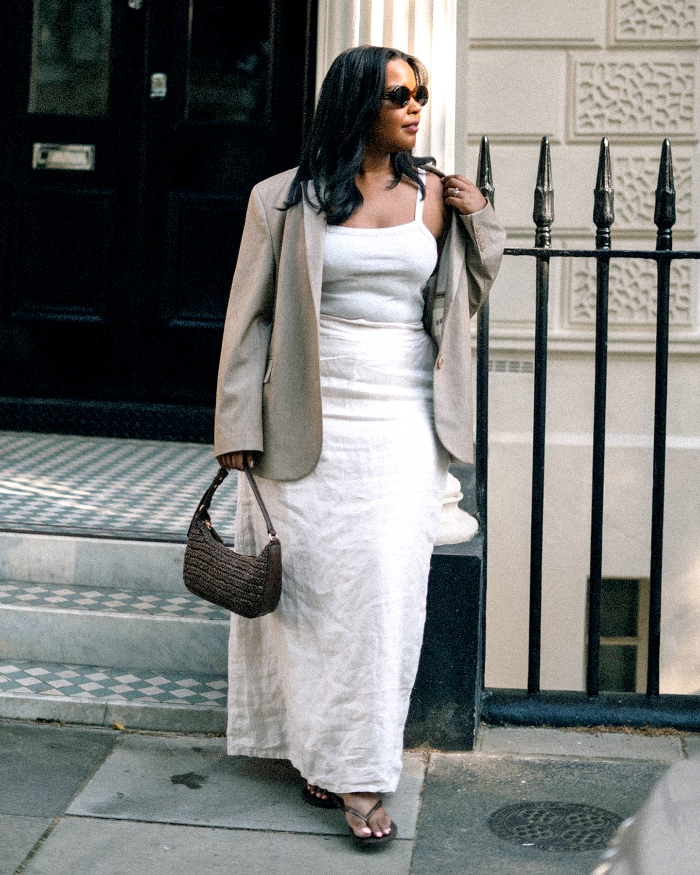 British fashion influencer Karina Marriott poses with oval sunglasses, a tan blazer, white sleeveless linen maxi dress, woven shoulder bag, and heeled flip-flop sandals