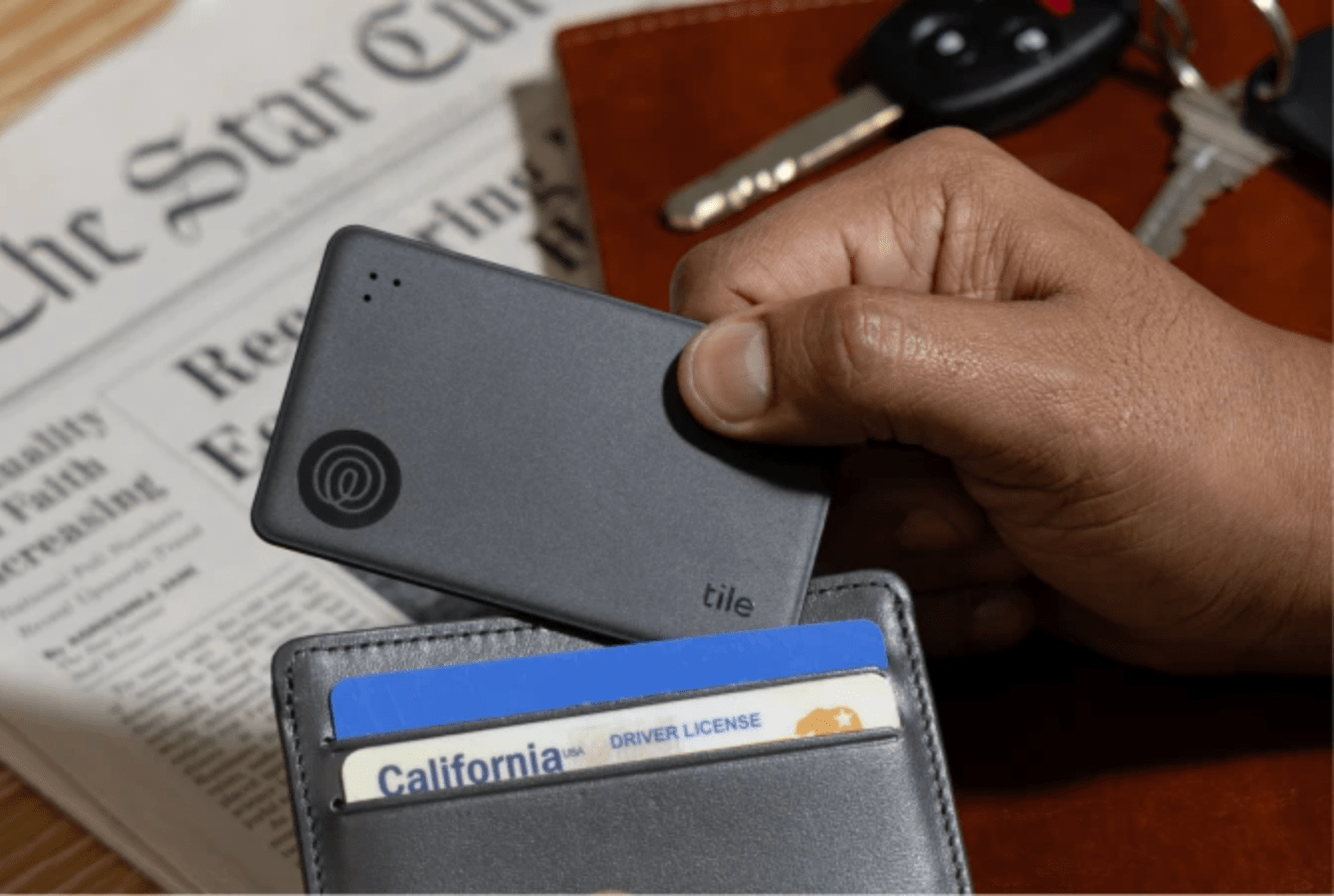 A hand placing a Tile Slim into a leather wallet with a newspaper in the background.