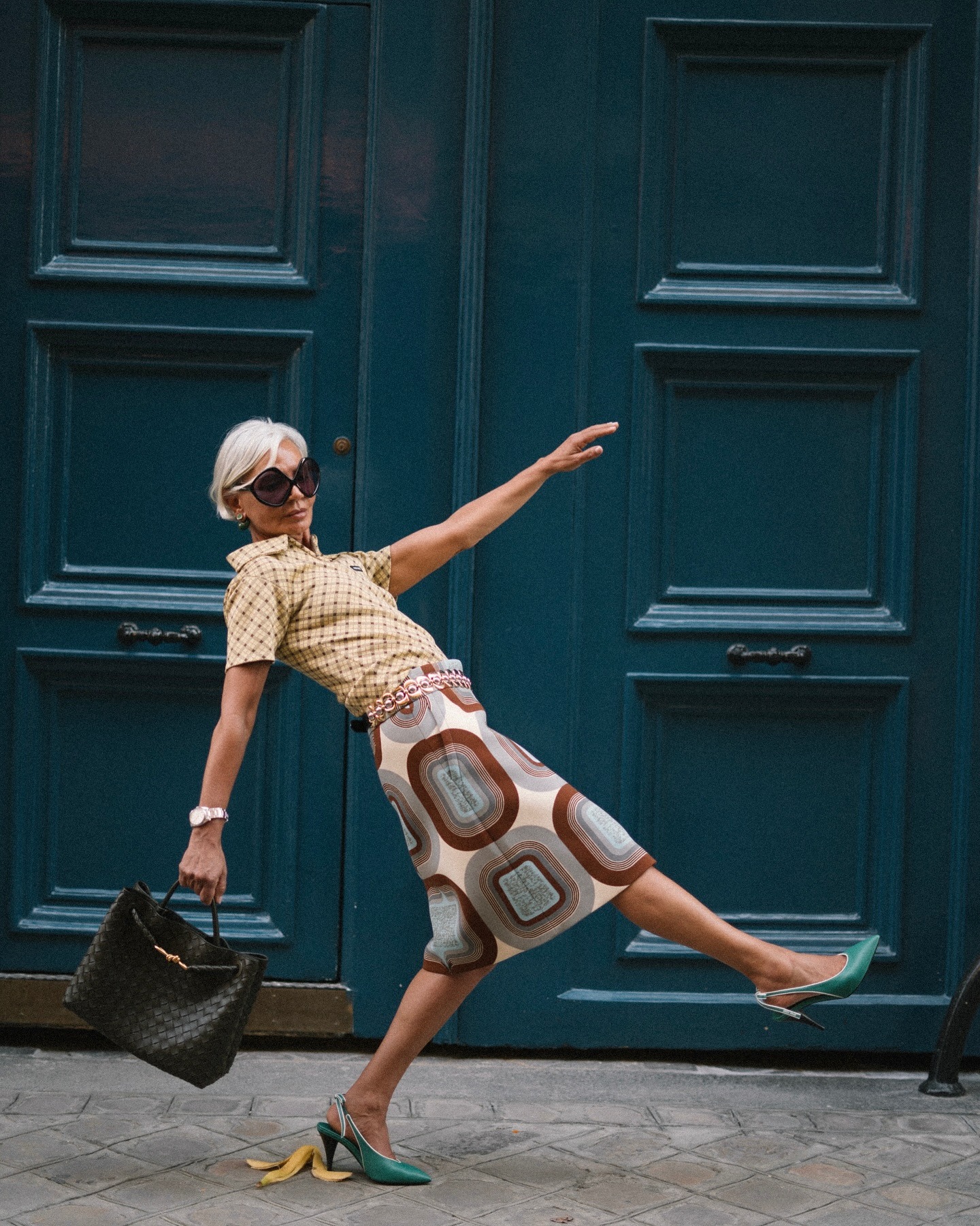 Grece wears a check short sleeve shirt, patterned midi skirt, blue kitten heels, and a bottega veneta tote bag.