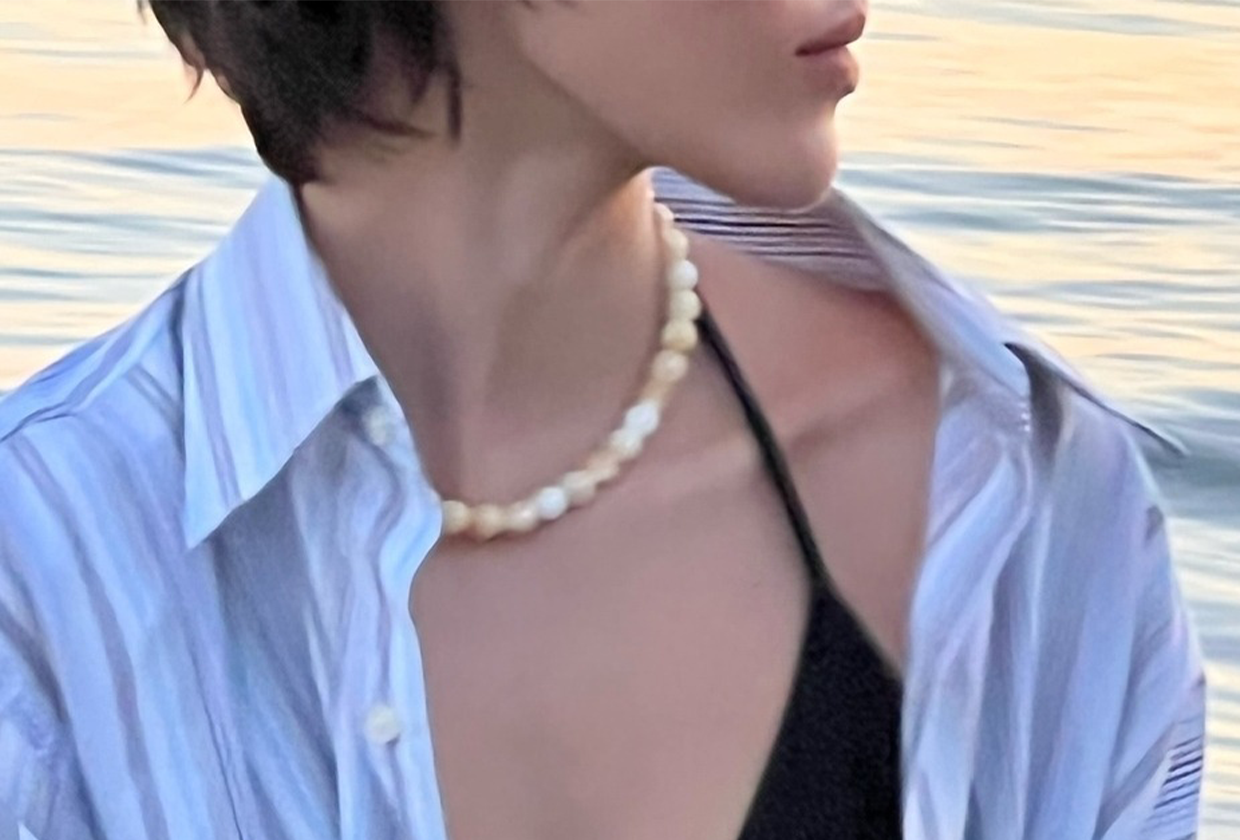 Model wearing black bikini, blue button-down shirt, and white stone necklace while at beach.