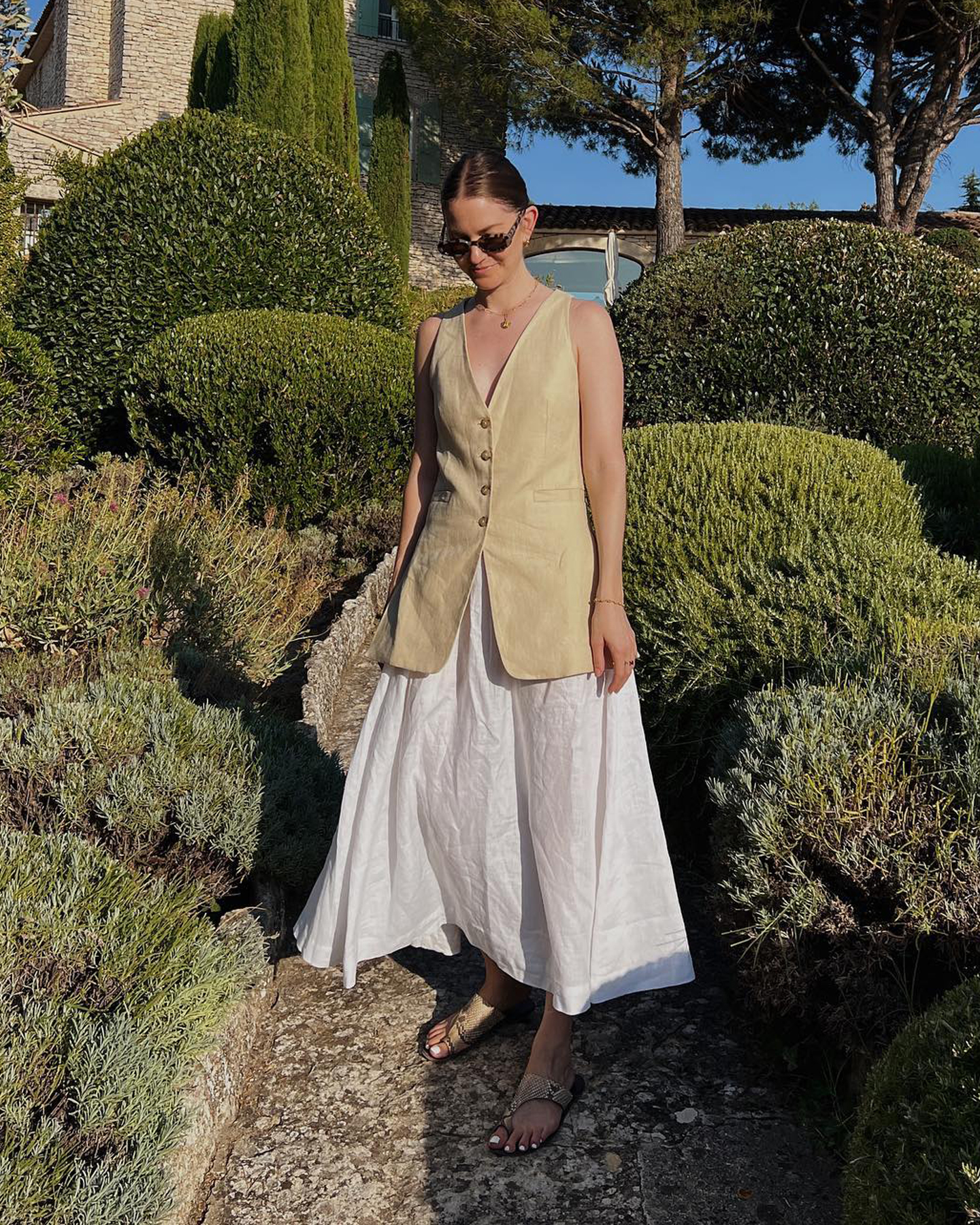 style influencer Marissa Cox poses in a lush French countryside garden wearing sunglasses, a tan long linen button-front vest, full white skirt, and toe ring snakeskin flat sandals