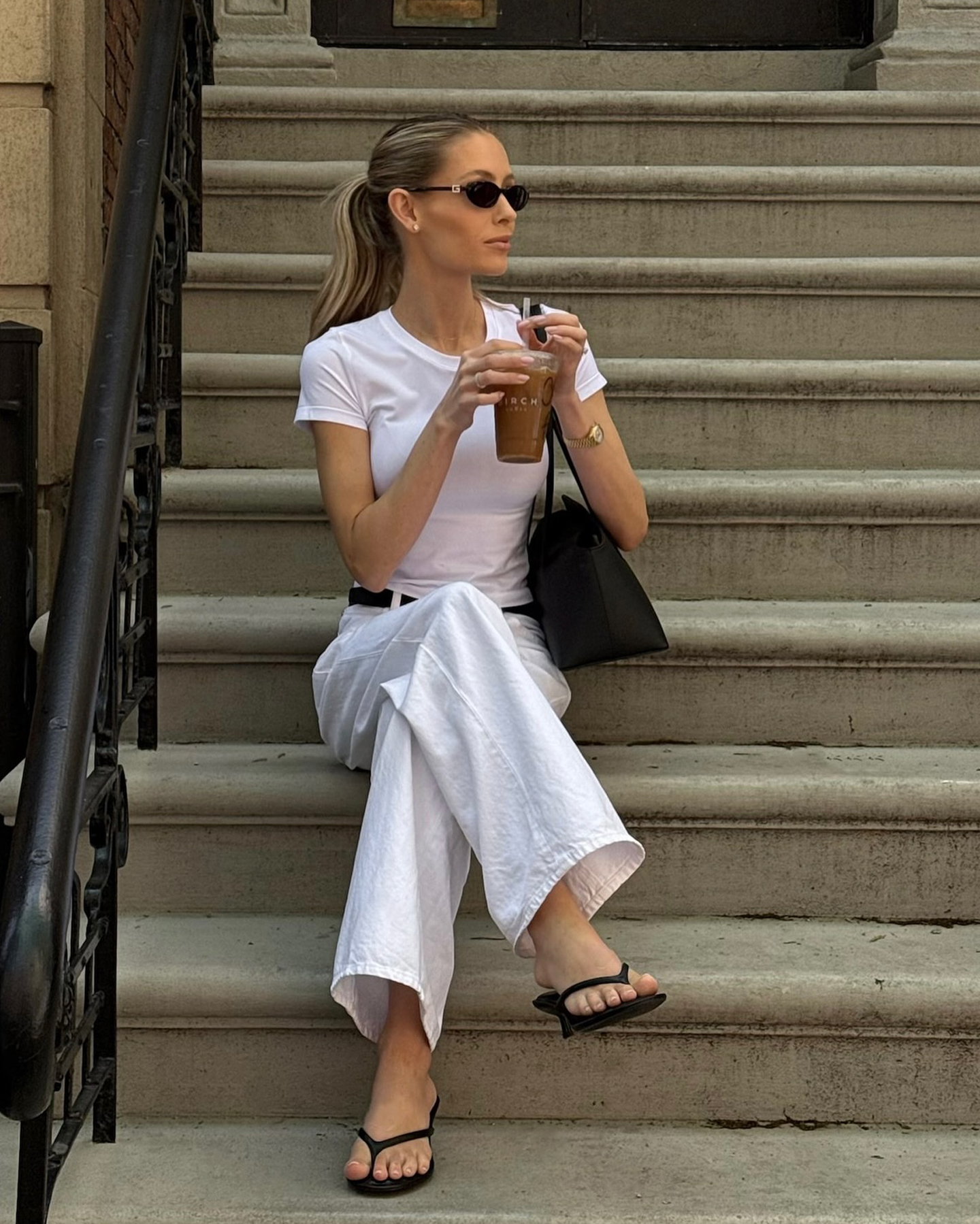 style influencer Fredrika Ekerot sits on a brownstone staircase in NYC with oval sunglasses, a white tee, iced coffee, leather shoulder bag, white jeans, and black heeled flip-flops